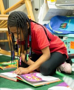 An elementary school-aged girl sits on the ground with a book in front of her, pointing at the words as she reads.